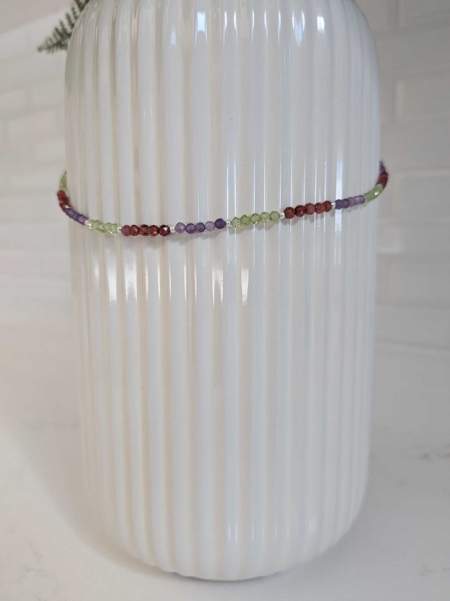 A delicate beaded bracelet with multicolored round beads, wrapped around a white ribbed ceramic vase.