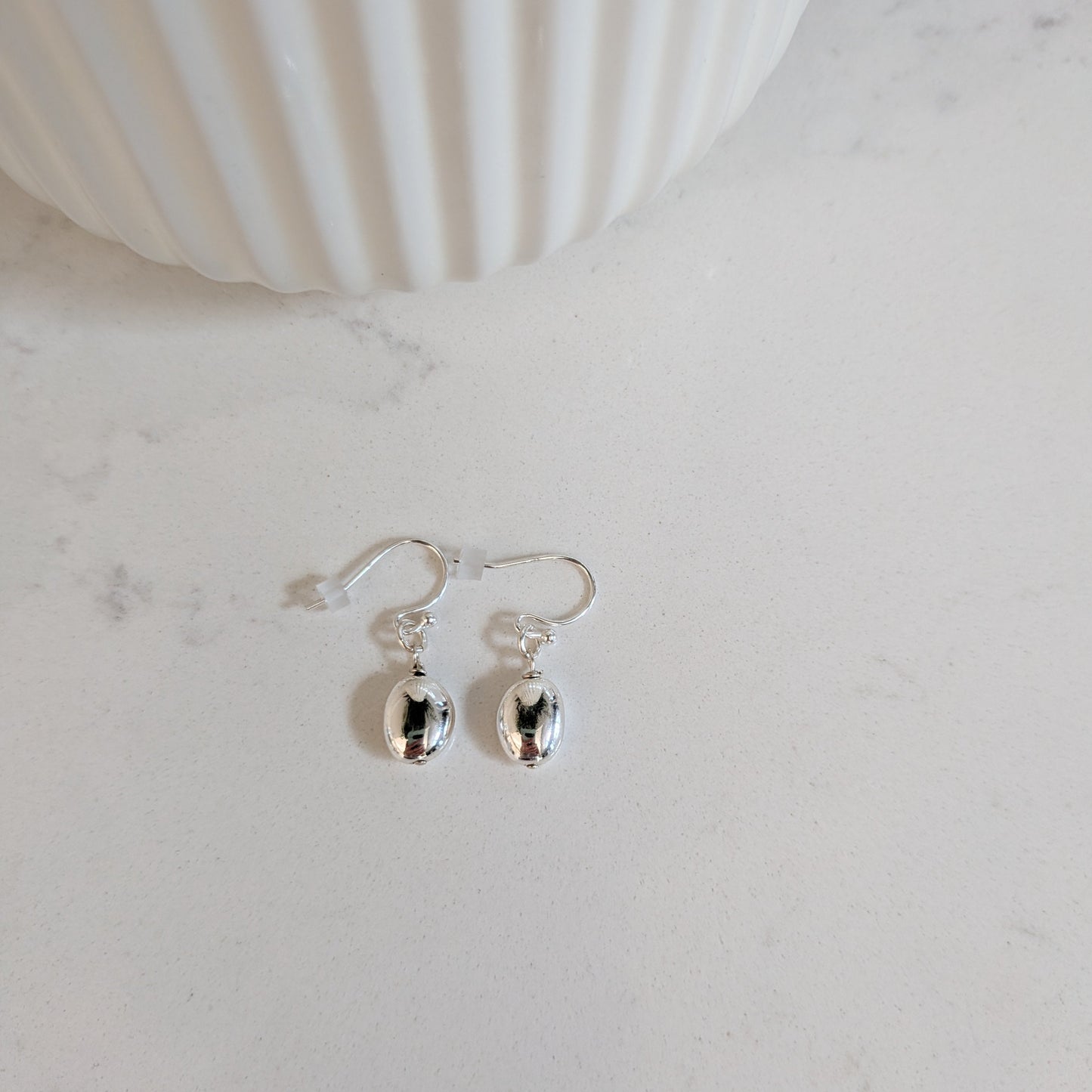 A pair of silver puffed oval drop earrings displayed on a light marble surface next to a white ribbed ceramic container.