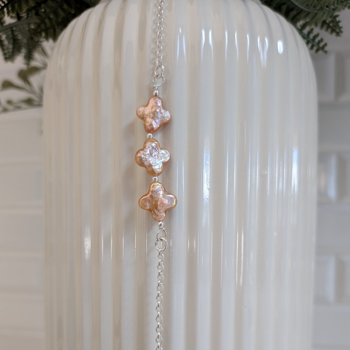 A silver rolo chain bracelet with three vertical champagne four leaf clover-pearls, hanging against a white ribbed ceramic vase.