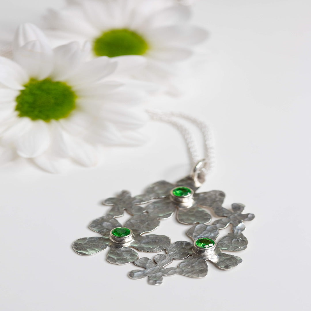Image of a Sterling Silver Handmade Daisy Delight Necklace adorned with vibrant tsavorite garnets. This stunning piece features a 20-gauge sterling silver pendant, tube-set tsavorite garnets, and a sterling silver wheat chain with a lobster clasp. Inspired by nature, the delicate daisy cluster design and captivating gemstones evoke a sense of beauty and transformation, perfect for reconnecting with nature's allure and awakening the beauty within.