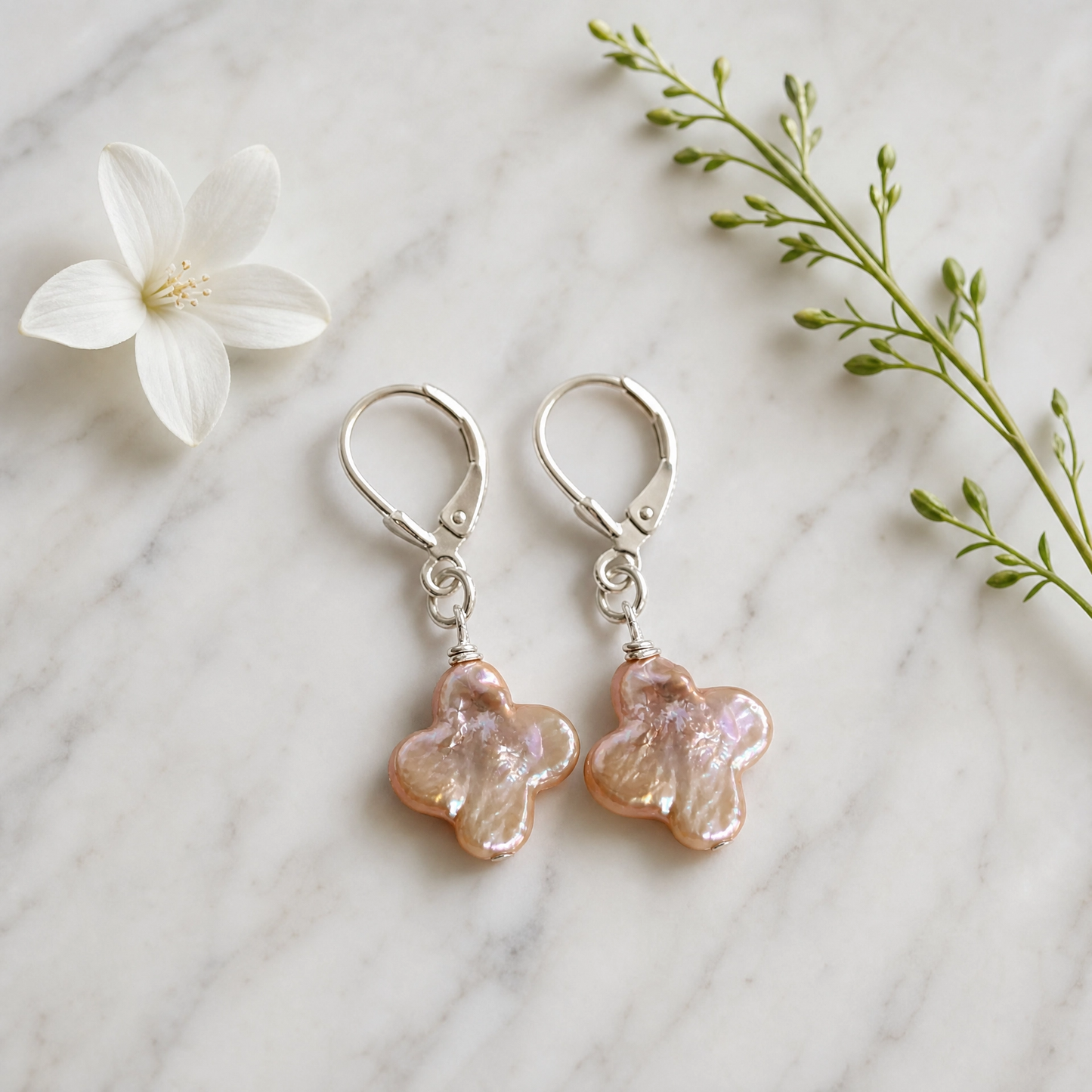 A pair of dangling earrings with silver leverback hooks and iridescent peach-pink flower-shaped charms, arranged on a white marble surface with a white flower and green sprig as props.