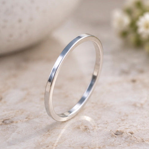 A polished silver band ring standing upright on a light beige stone surface, with a softly blurred white ceramic object and small white flowers in the background.