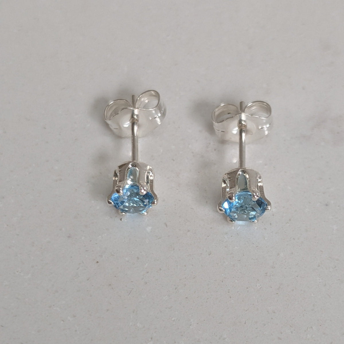A pair of silver stud earrings with small light blue gemstones, shown on a light gray textured surface.