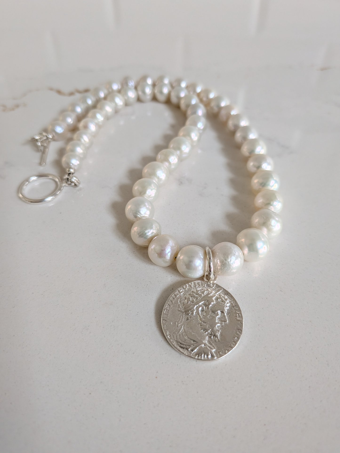 A pearl necklace with a silver coin pendant, displayed on a light marble surface. The necklace features white freshwater-style pearls with a silver-tone clasp and a circular silver coin charm.
