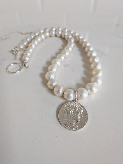 A pearl necklace with a silver coin pendant, displayed on a light marble surface. The necklace features white freshwater-style pearls with a silver-tone clasp and a circular silver coin charm.