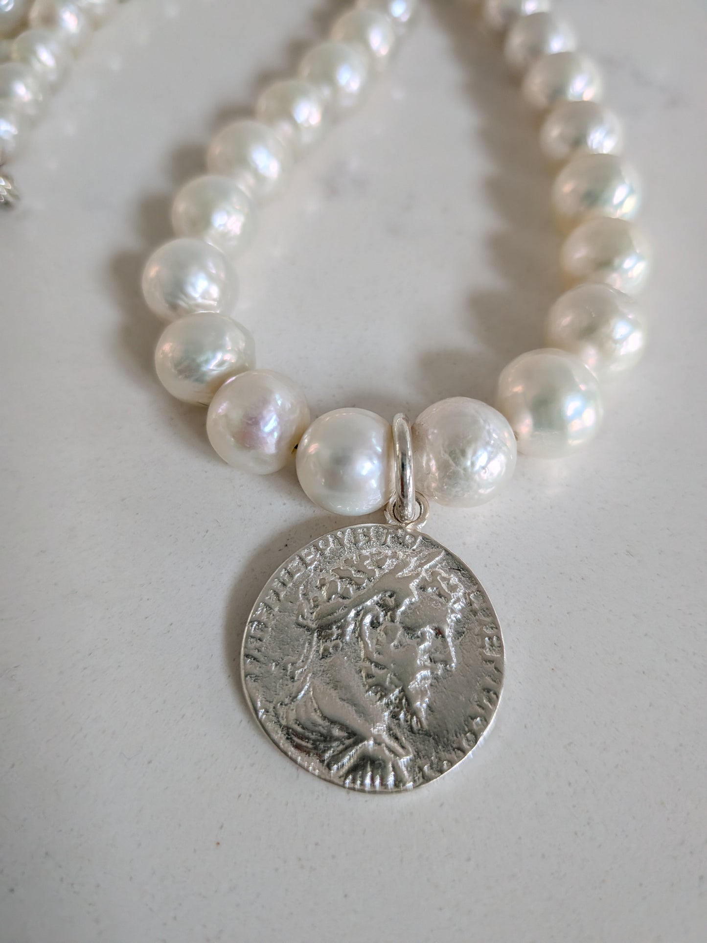 A close-up of a beaded necklace with white pearl-like round beads, a small silver connector, and a dangling silver coin-style pendant showing an embossed profile portrait.