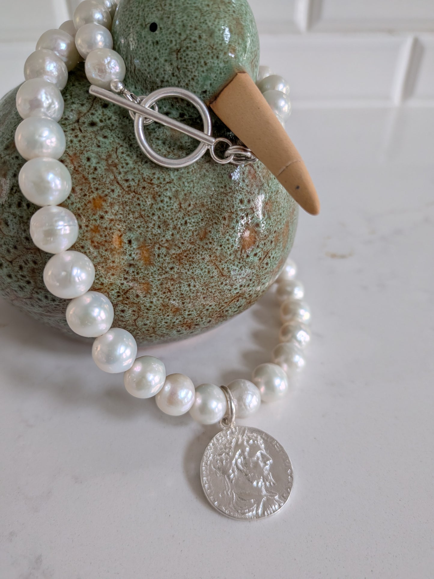 A beaded pearl necklace with a silver toggle clasp and a silver coin pendant, displayed on a green ceramic duck figurine against a white tiled background.