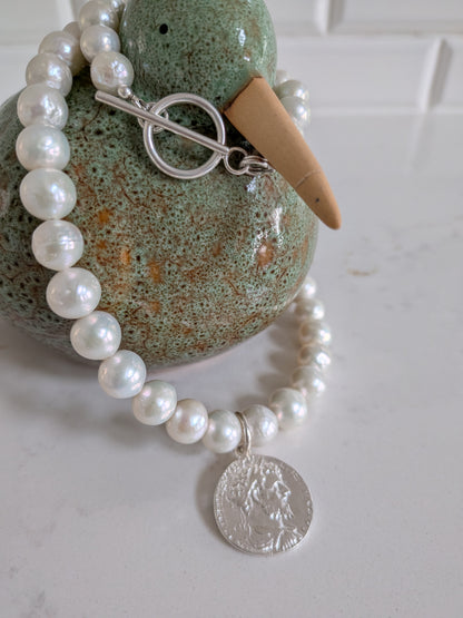 A beaded pearl necklace with a silver toggle clasp and a silver coin pendant, displayed on a green ceramic duck figurine against a white tiled background.