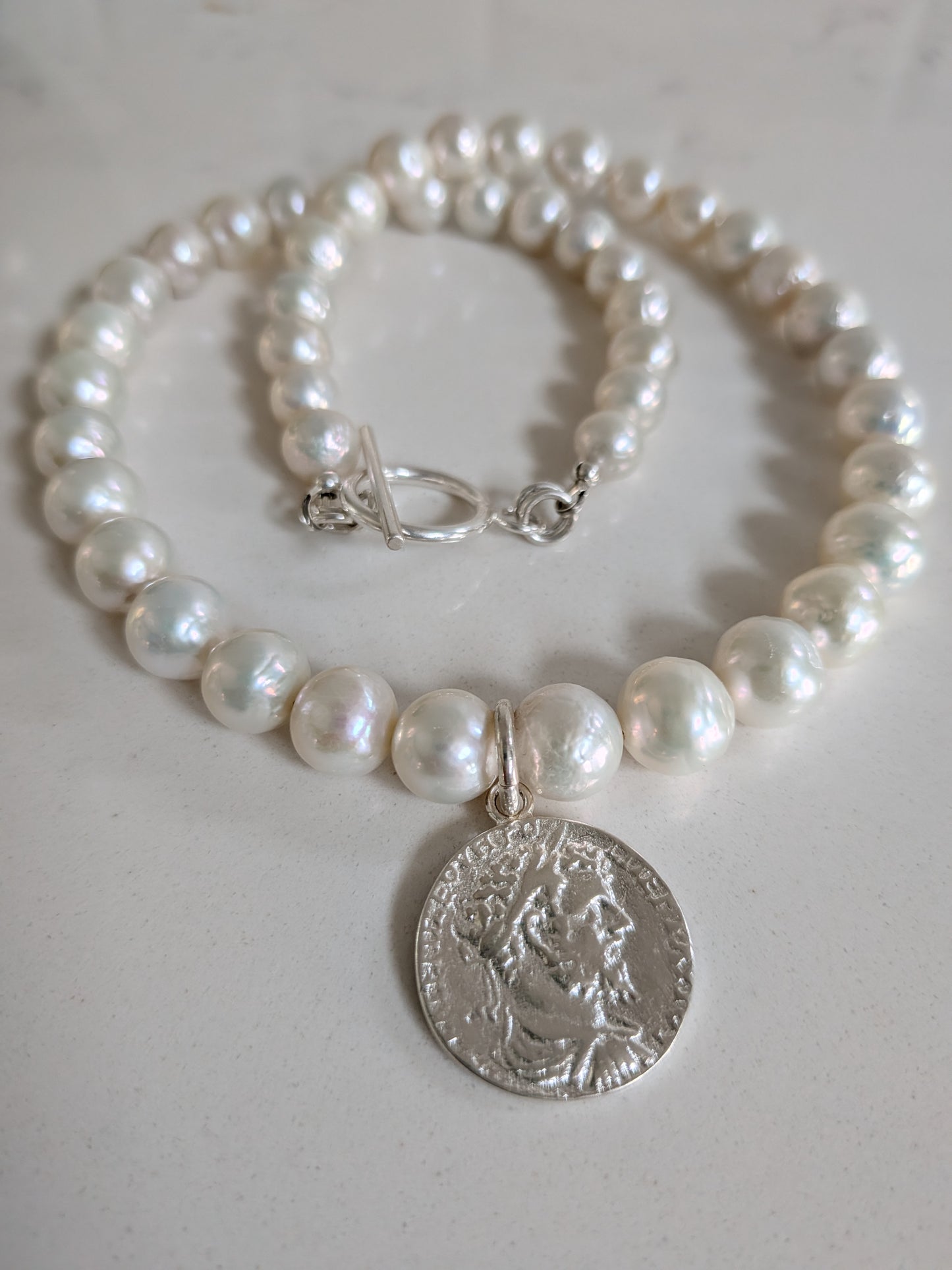 A pearl beaded necklace with a silver-toned coin pendant featuring a classical profile, displayed on a light neutral surface.