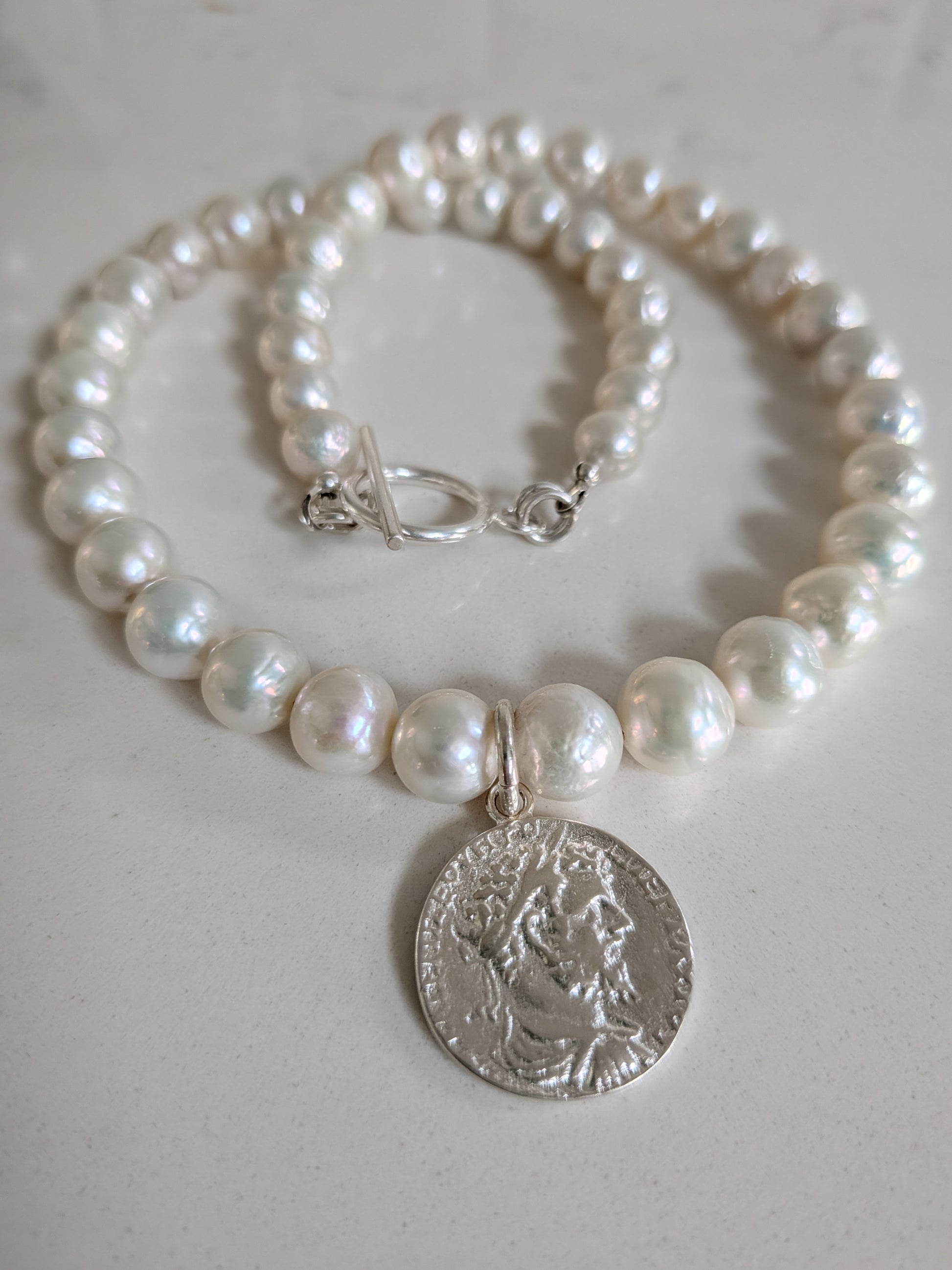 A pearl beaded necklace with a silver-toned coin pendant featuring a classical profile, displayed on a light neutral surface.