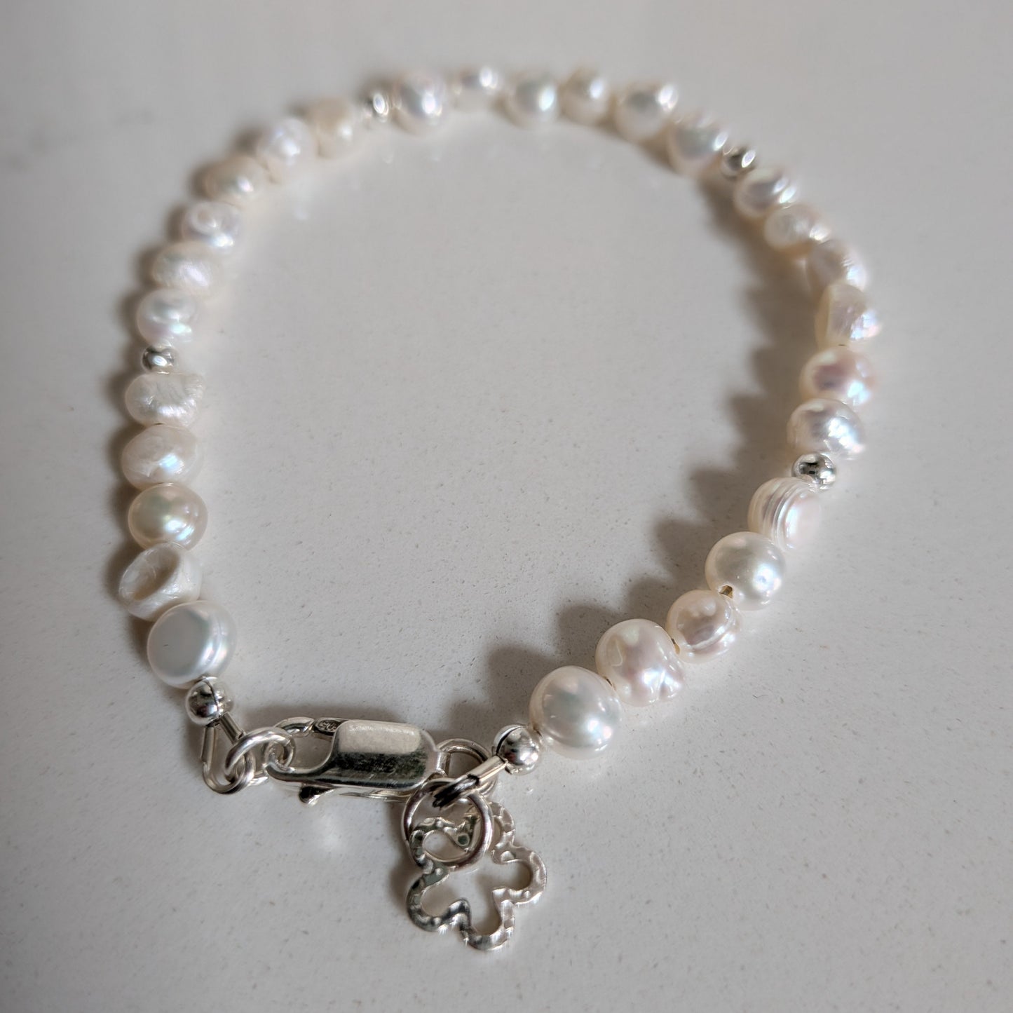 A freshwater pearl bracelet with irregular white pearls and a silver lobster clasp featuring a small dangling flower silver charm, displayed on a light gray surface.