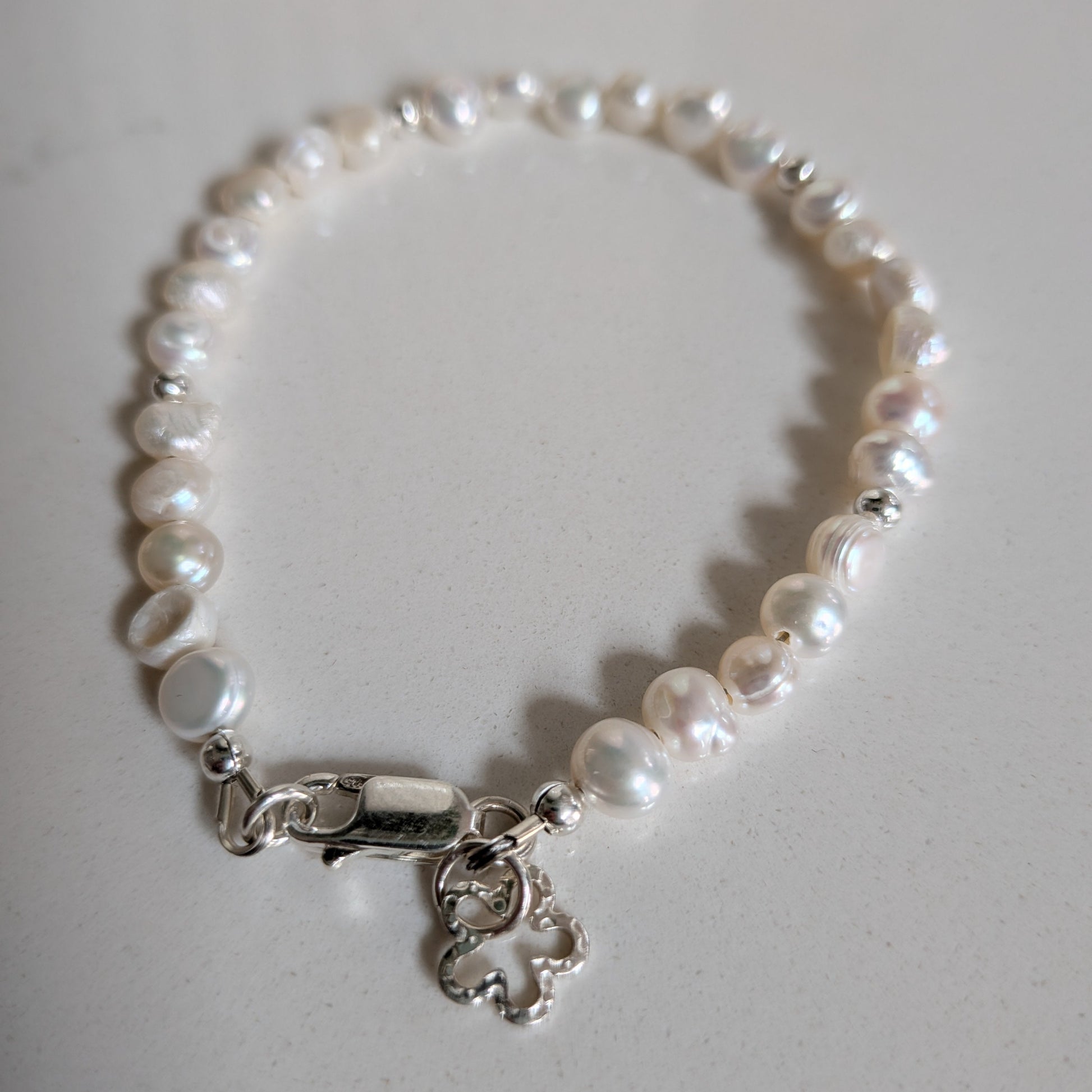 A freshwater pearl bracelet with irregular white pearls and a silver lobster clasp featuring a small dangling flower silver charm, displayed on a light gray surface.