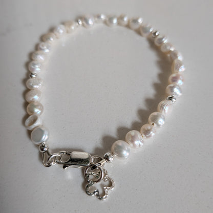A freshwater pearl bracelet with irregular white pearls and a silver lobster clasp featuring a small dangling flower silver charm, displayed on a light gray surface.