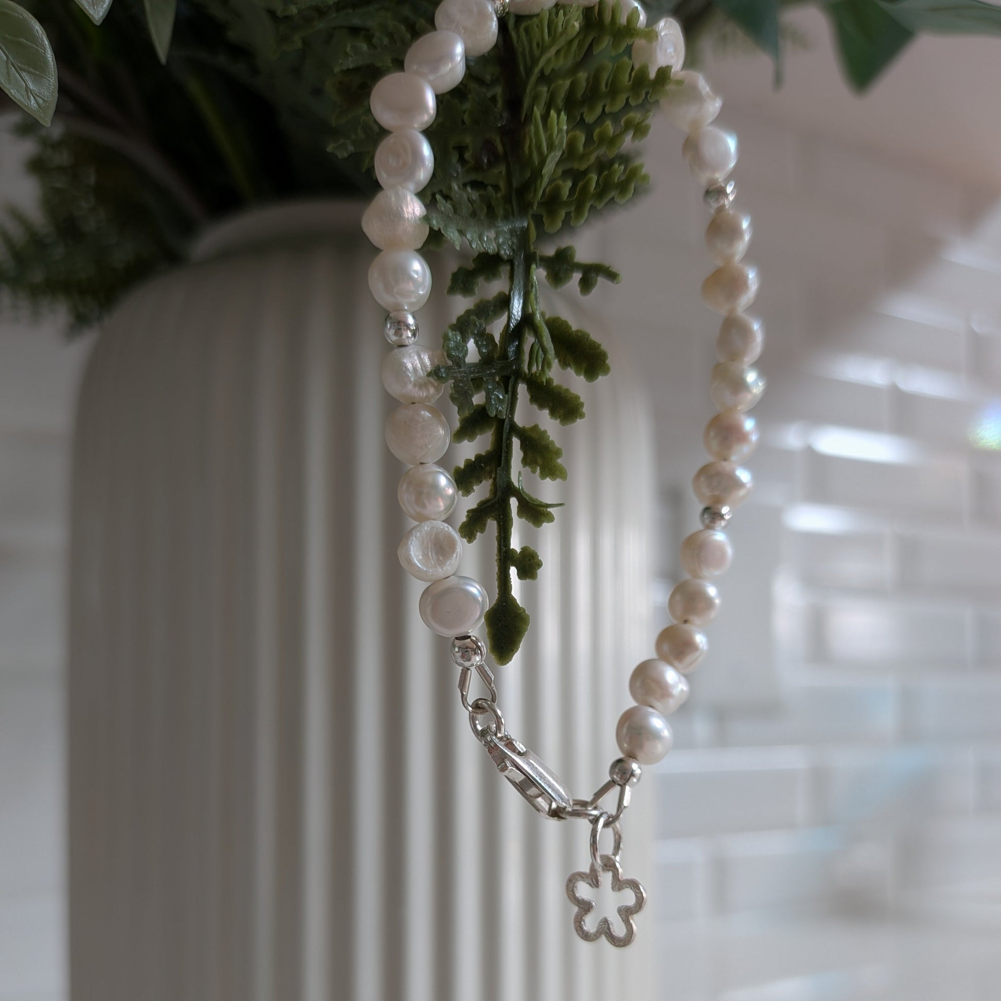 A strand of white pearl-like beads with a silver clasp and a small flower-shaped charm, draped over a white ribbed vase with green foliage in the background.