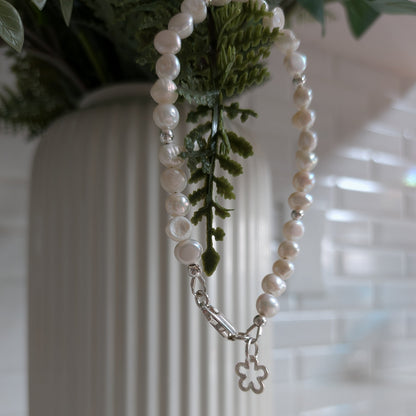 A strand of white pearl-like beads with a silver clasp and a small flower-shaped charm, draped over a white ribbed vase with green foliage in the background.