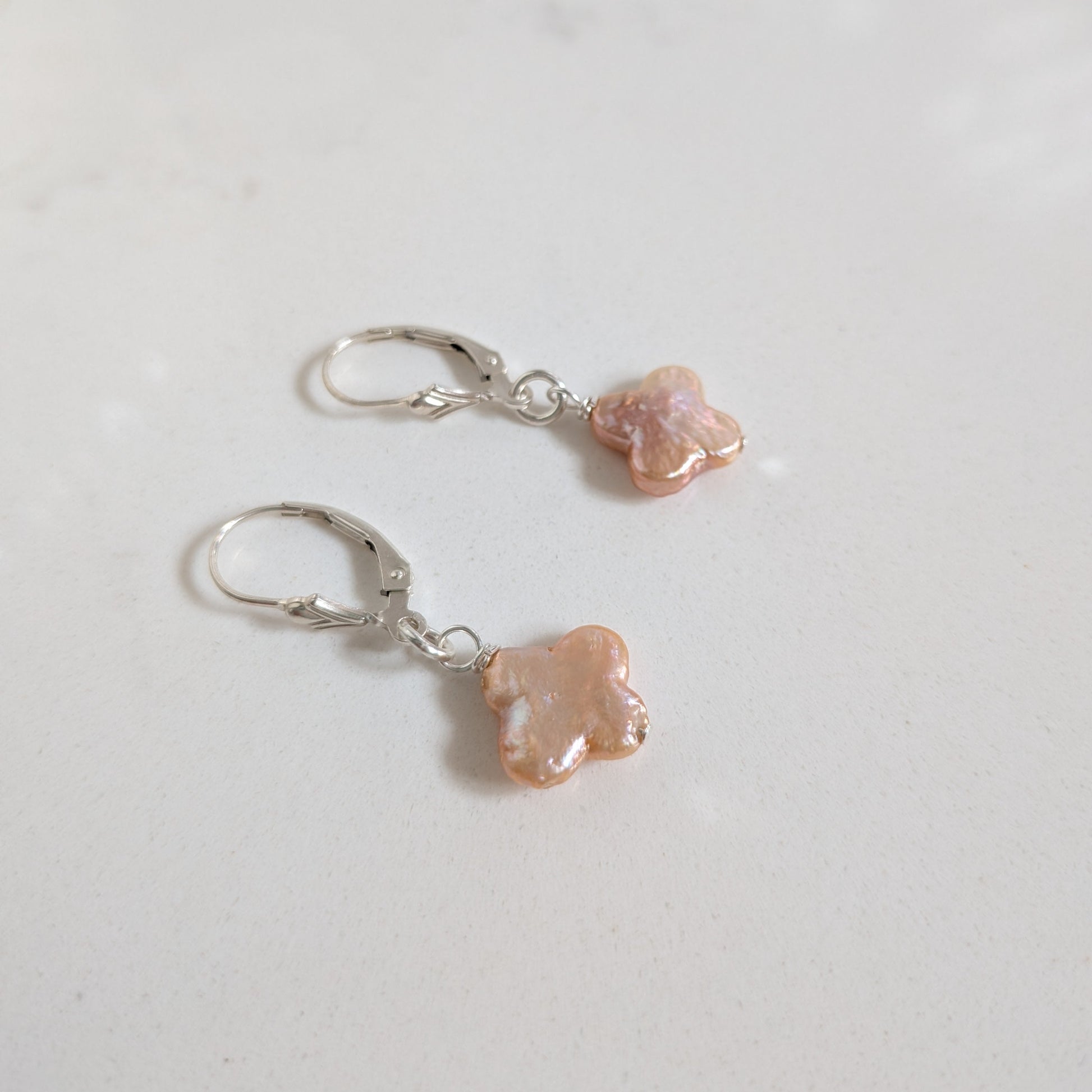 A pair of silver leverback earrings with small, irregularly shaped peach-pink beads, displayed on a light gray surface.