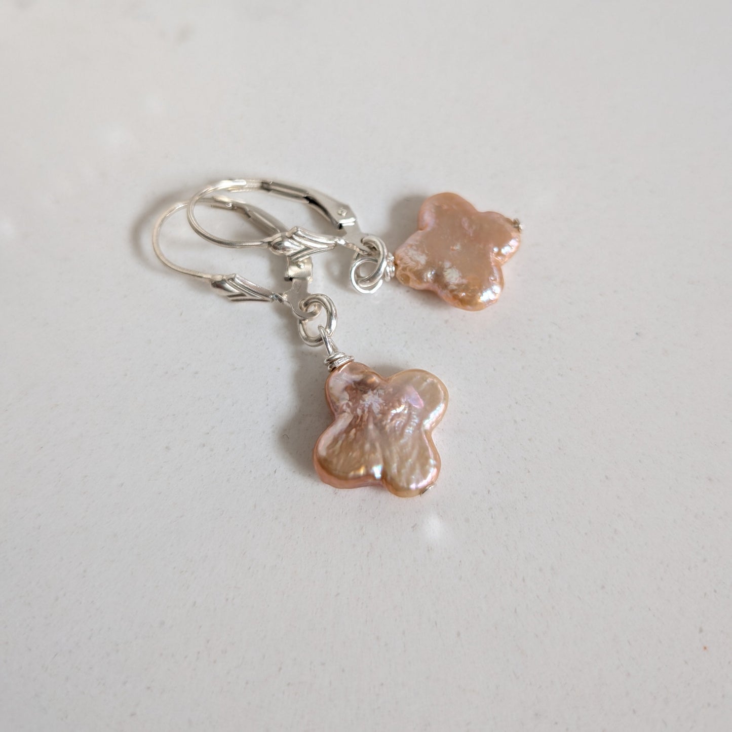 A pair of silver leverback earrings with irregular, pearlescent peach-pink flower-shaped charms, displayed on a light gray surface.