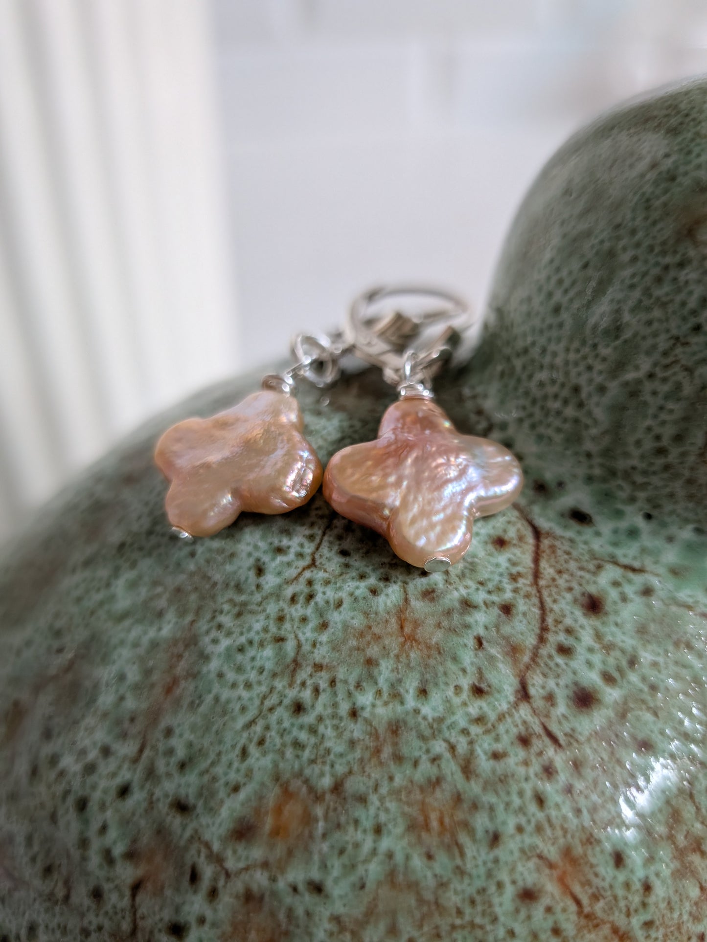 A pair of handmade dangle earrings with irregular, flower-like peach and pink stone pendants on silver leverbacks, resting on a textured green ceramic surface.
