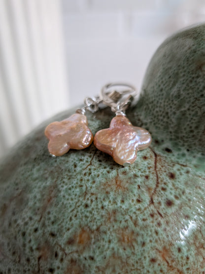 A pair of handmade dangle earrings with irregular, flower-like peach and pink stone pendants on silver leverbacks, resting on a textured green ceramic surface.