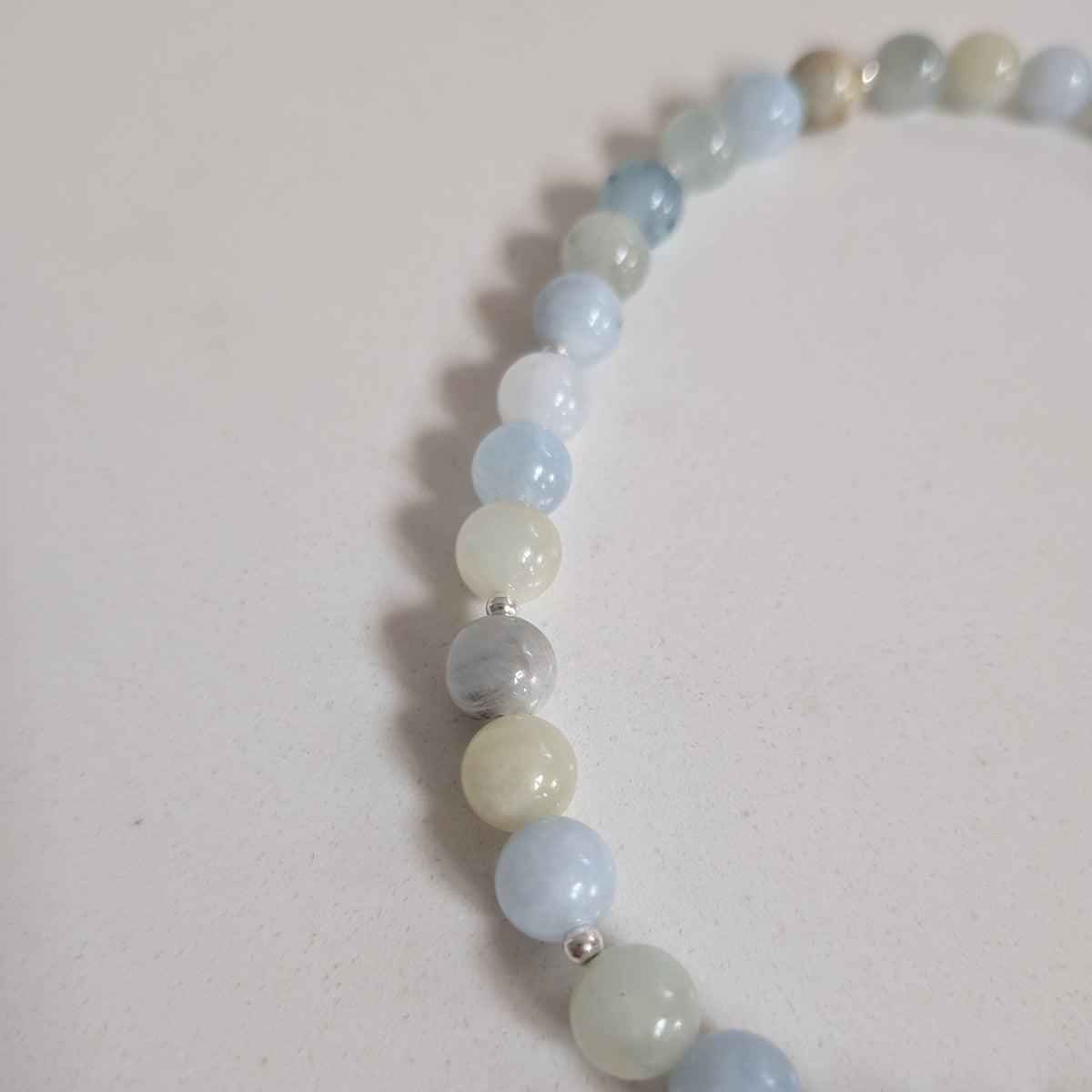 A beaded stretch bracelet made of smooth, polished round gemstone beads in light blue, pale green, white, and gray, with small silver spacer beads, shown on a neutral off-white background.