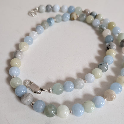 A beaded necklace made of polished round stones in light blue, pale green, white, and gray, with small silver spacer beads and a silver lobster clasp, photographed on a light marble-like surface.