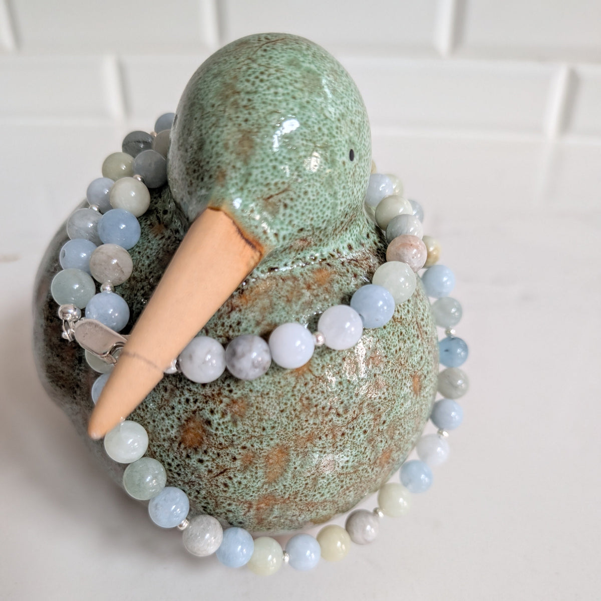 A handmade ceramic duck figurine with a speckled green glaze and a natural wood beak, wrapped in a beaded necklace of light blue, white, and gray round beads, set against a white tiled background.