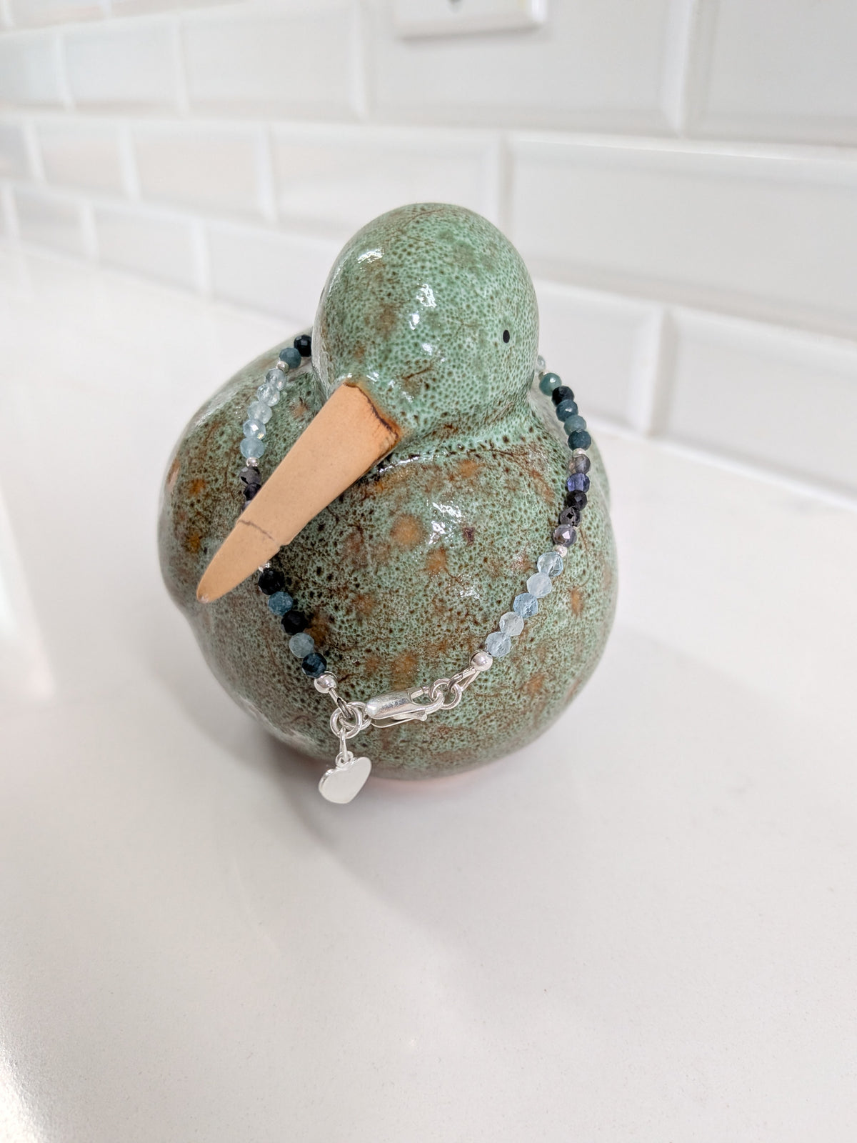 A handmade ceramic duck figurine with a speckled green and brown glaze wearing a beaded bracelet with a silver heart charm, set on a white countertop with a white subway tile backsplash.