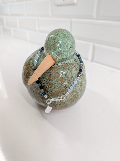 A handmade ceramic duck figurine with a speckled green and brown glaze wearing a beaded bracelet with a silver heart charm, set on a white countertop with a white subway tile backsplash.