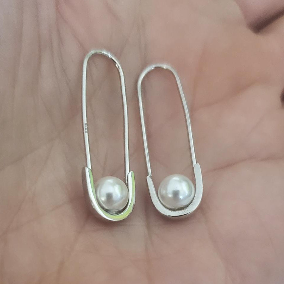 Two silver safety pins with embedded pearls held in a hand against a neutral background