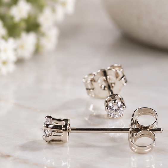 A pair of silver stud earrings with clear 3mm cubic zirconia, displayed on a white marble surface with soft-focus white flowers in the background.