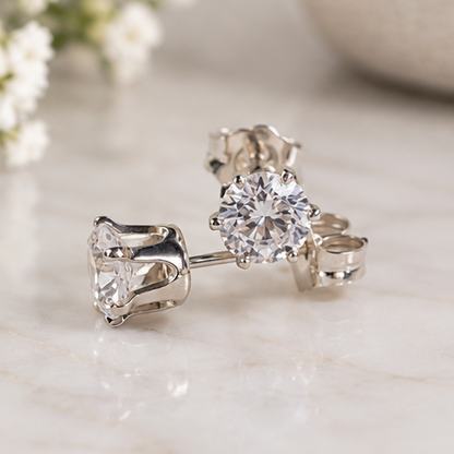 A pair of silver stud earrings with 5mm crystal cubic zirconias, displayed on a light marble surface with soft-focus flowers in the background.