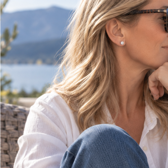 A close-up lifestyle shot of a woman with blonde hair wearing 5mm crystal cubic zirconia earring, a white linen shirt, and blue jeans, sitting outdoors with a scenic lake and mountains in the background.