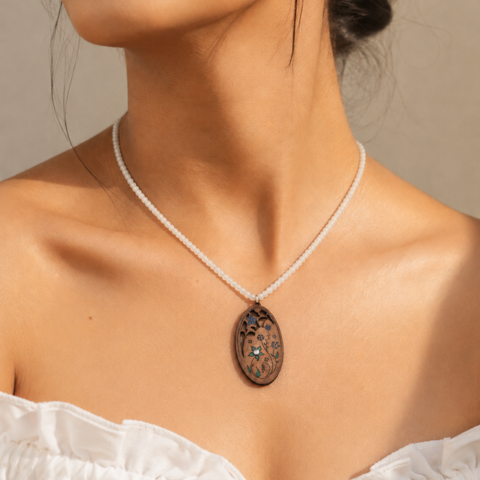 A close-up of a model wearing a delicate aquamarine beaded necklace with an oval leather pendant. The pendant features a dark brown background with a detailed floral pattern and small turquoise blue accents.
