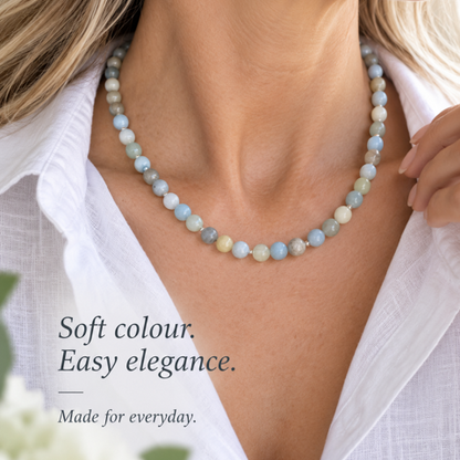 A close-up lifestyle shot of a woman wearing a beaded necklace made of light blue, mint green, and cream beads. She is wearing a white linen shirt, and the text on the left reads 'Soft colour.