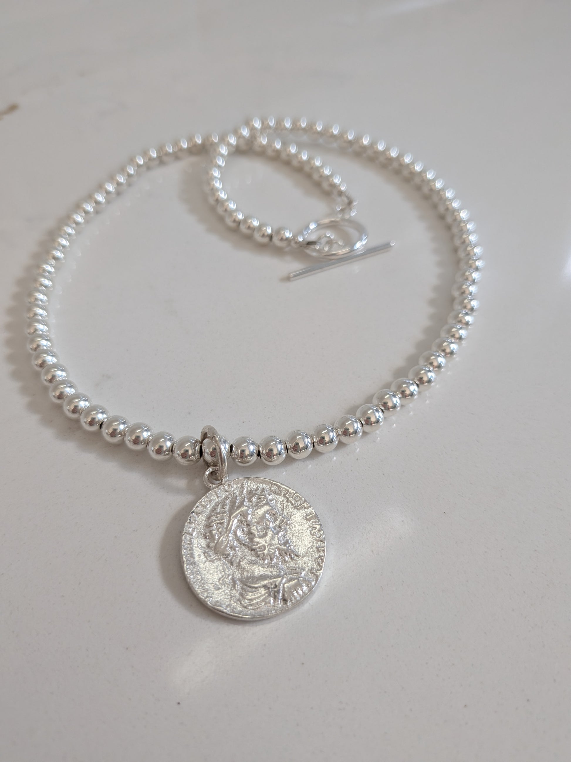 A silver beaded bracelet with a round coin pendant and toggle clasp, displayed on a light off-white surface.