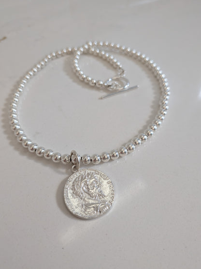 A silver beaded bracelet with a round coin pendant and toggle clasp, displayed on a light off-white surface.