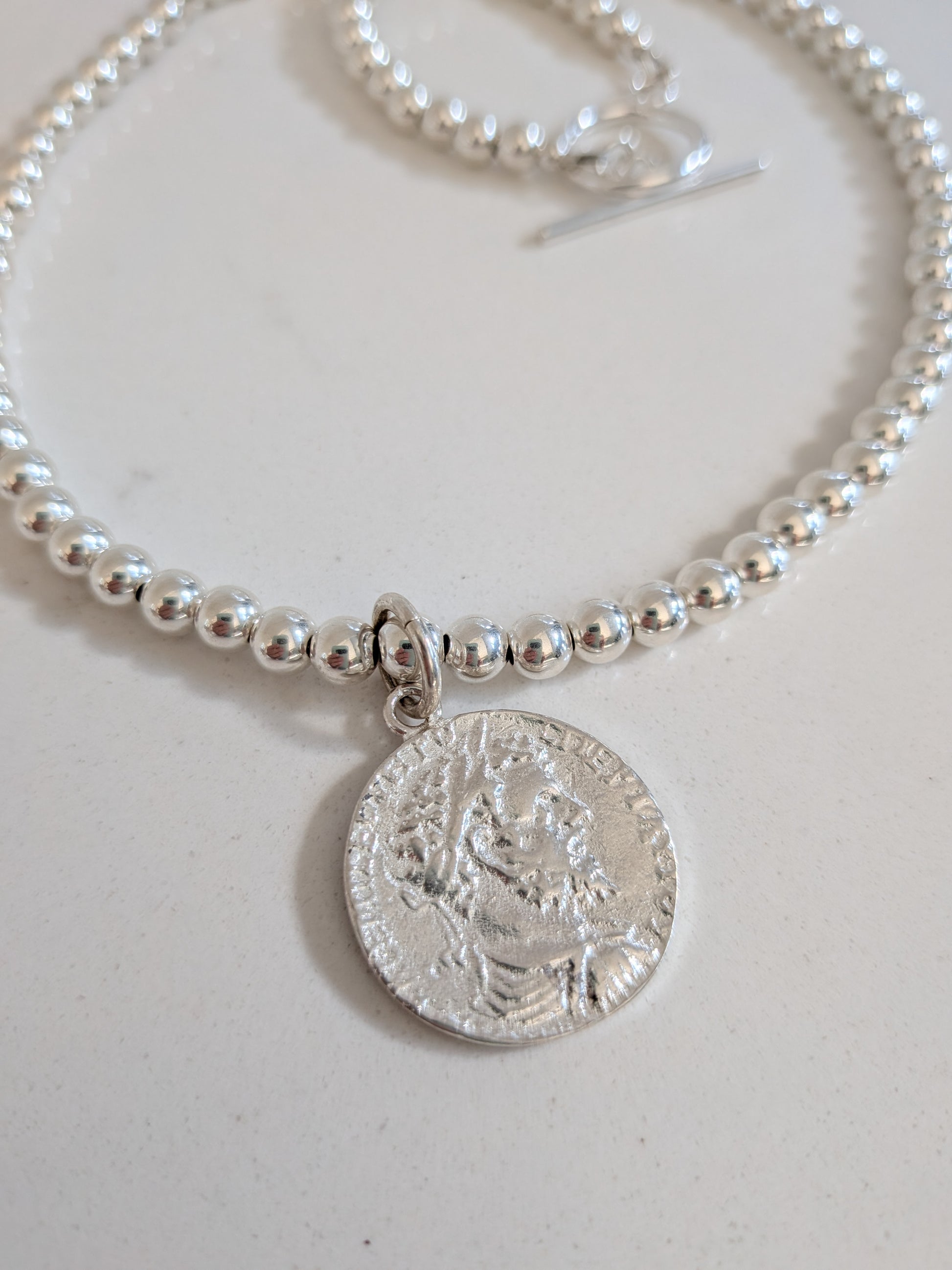 A silver beaded necklace with a circular coin-style pendant featuring an embossed portrait, displayed on a light off-white surface.