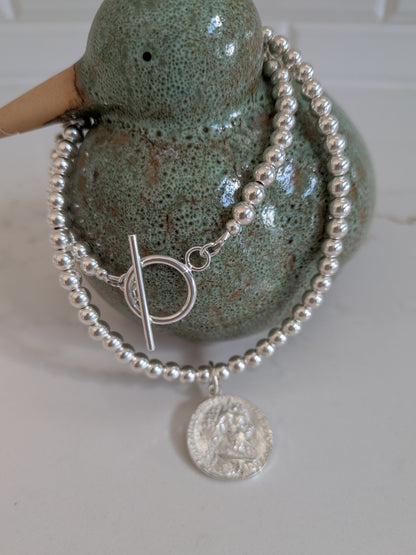 A sterling silver beaded bracelet with a toggle clasp and a round coin pendant, draped over a green ceramic duck-shaped display holder.