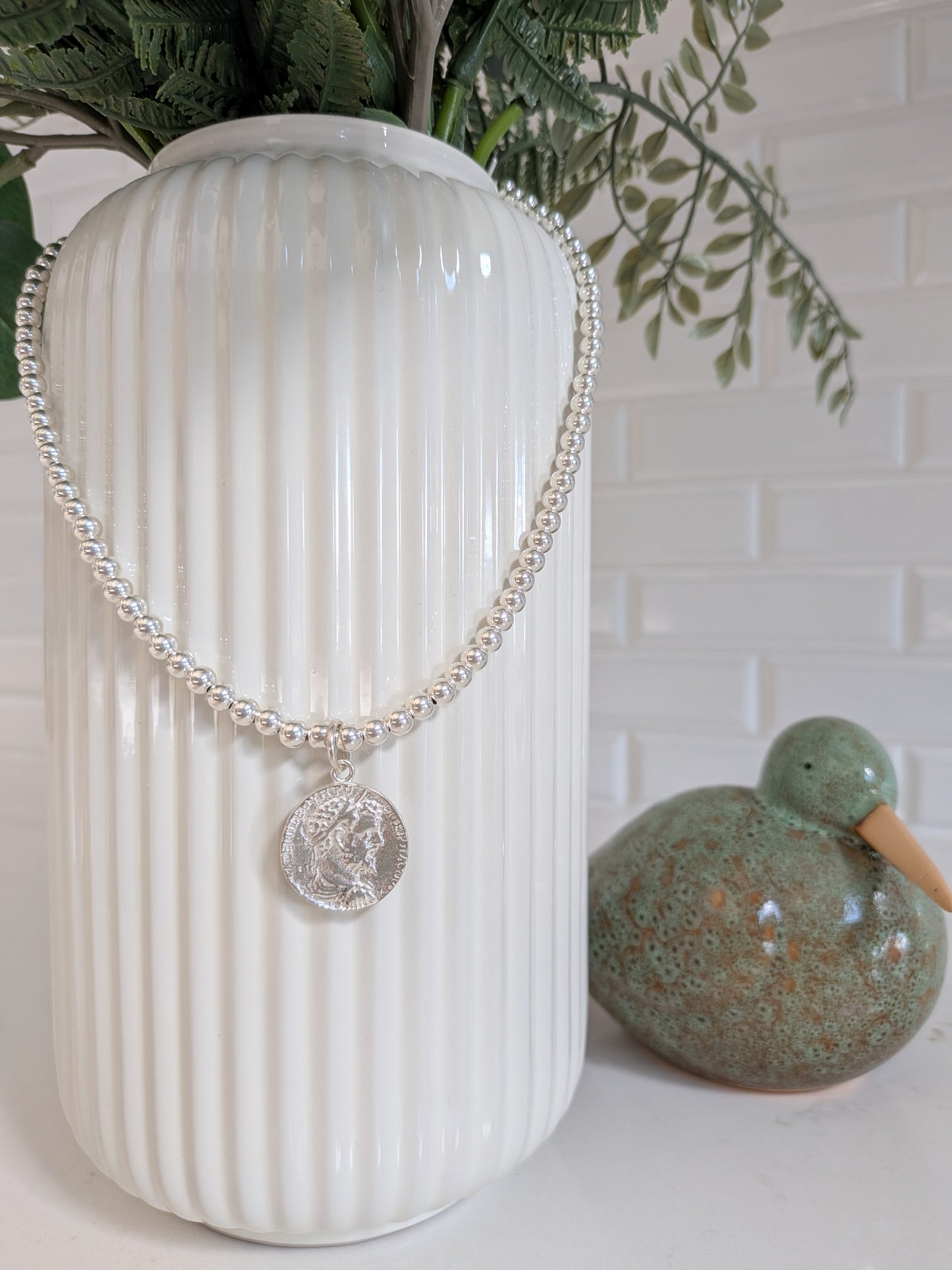 A silver beaded necklace with a circular coin pendant displayed on a white ribbed ceramic vase, with green foliage and a green duck figurine in the background.