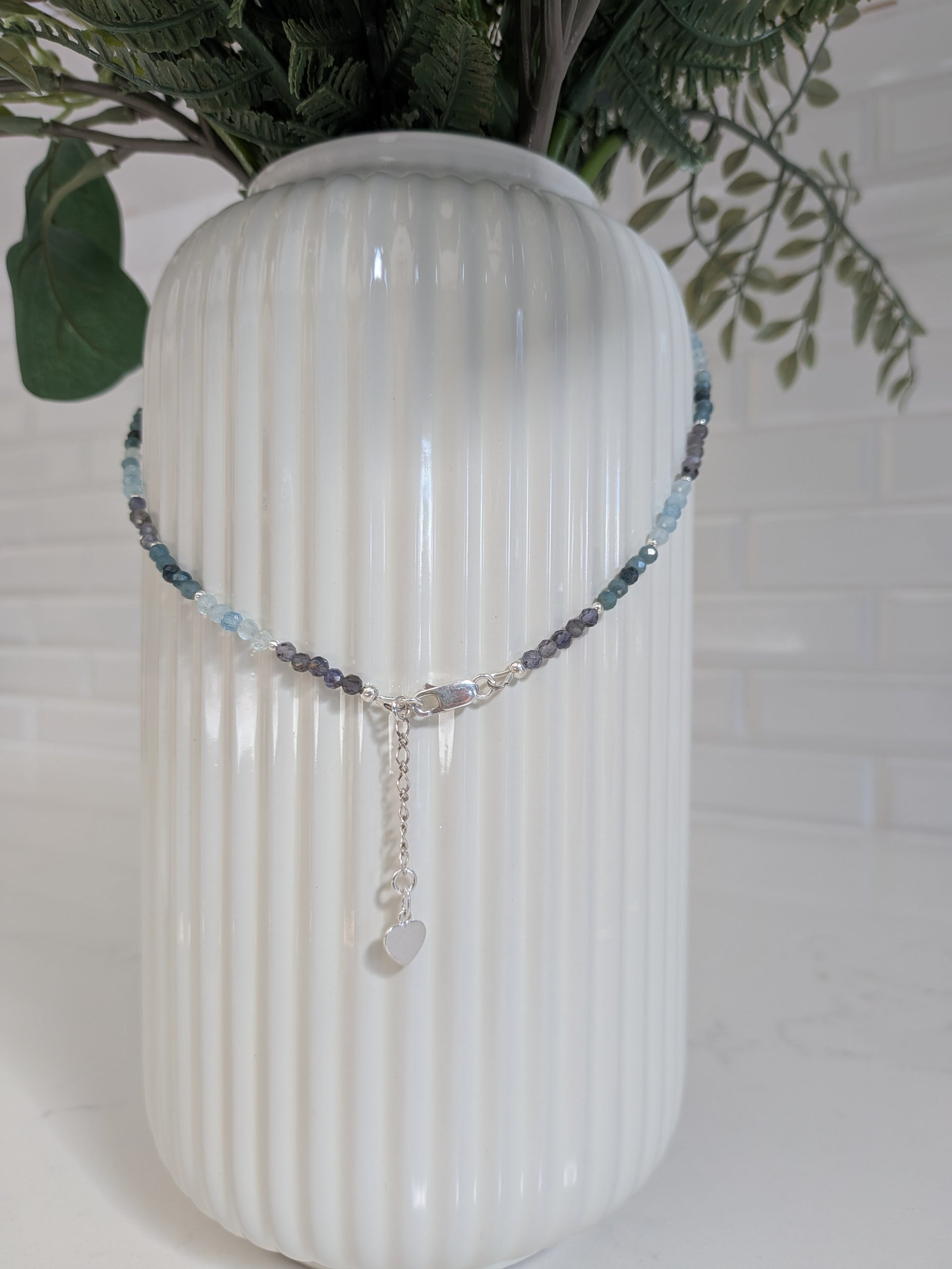 A beaded necklace with light blue, gray, and dark purple beads, featuring a silver-tone clasp and a small heart-shaped charm, draped over a white ribbed ceramic vase.