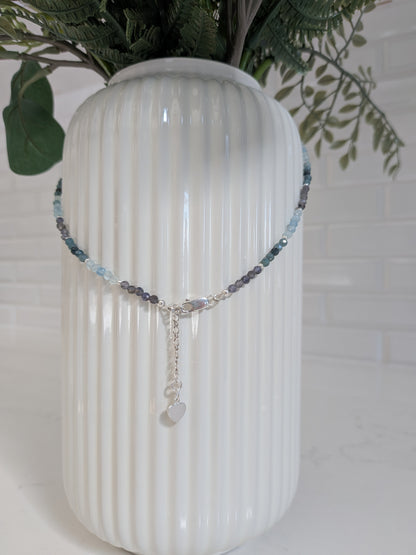 A beaded necklace with light blue, gray, and dark purple beads, featuring a silver-tone clasp and a small heart-shaped charm, draped over a white ribbed ceramic vase.