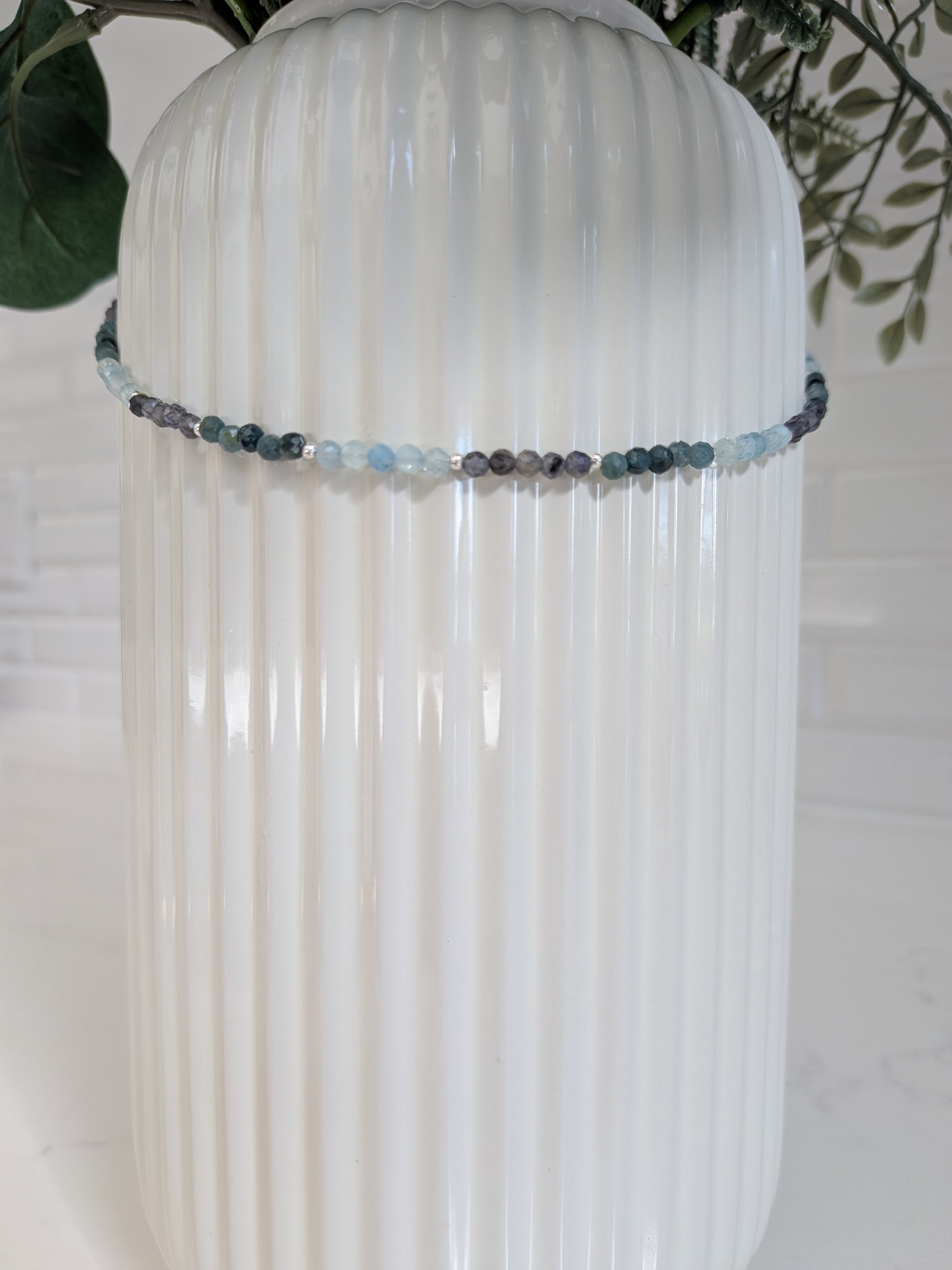 A beaded crystal necklace with light blue, gray, and dark charcoal beads draped around a white ribbed ceramic vase.
