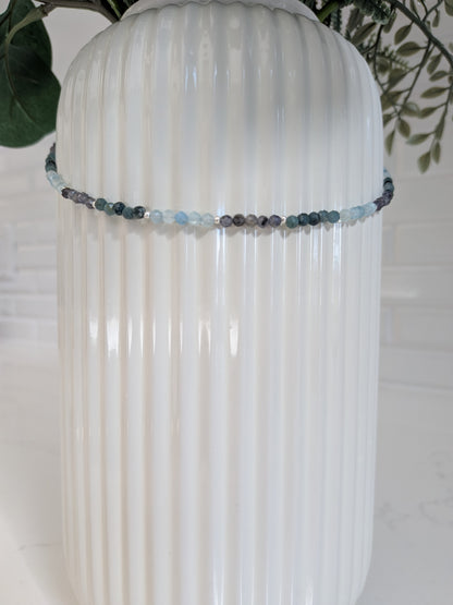A beaded crystal necklace with light blue, gray, and dark charcoal beads draped around a white ribbed ceramic vase.