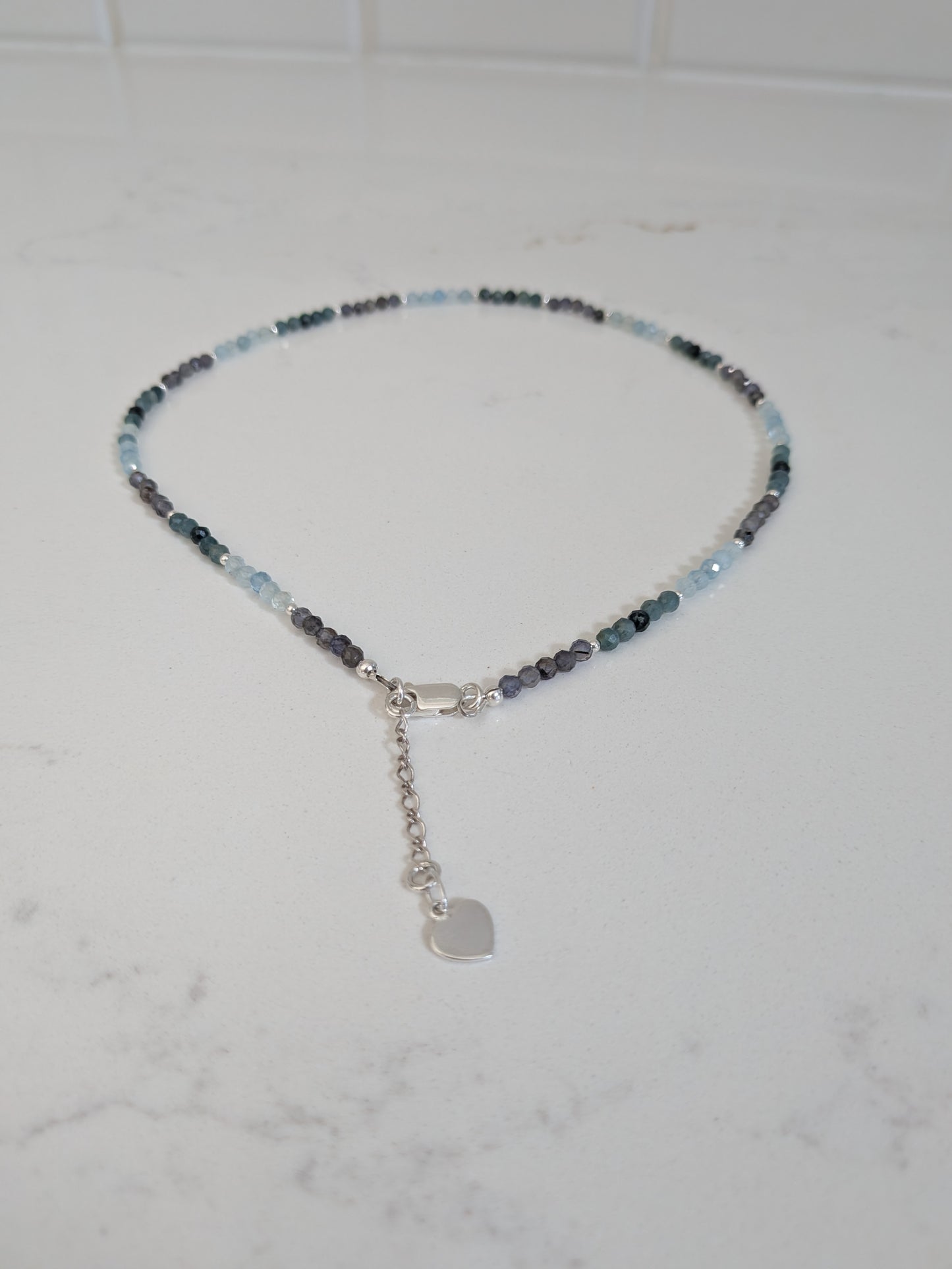 A delicate beaded necklace with small light blue, dark blue, and gray beads, featuring a silver lobster clasp, extender chain, and a small heart-shaped charm, displayed on a white marble surface.
