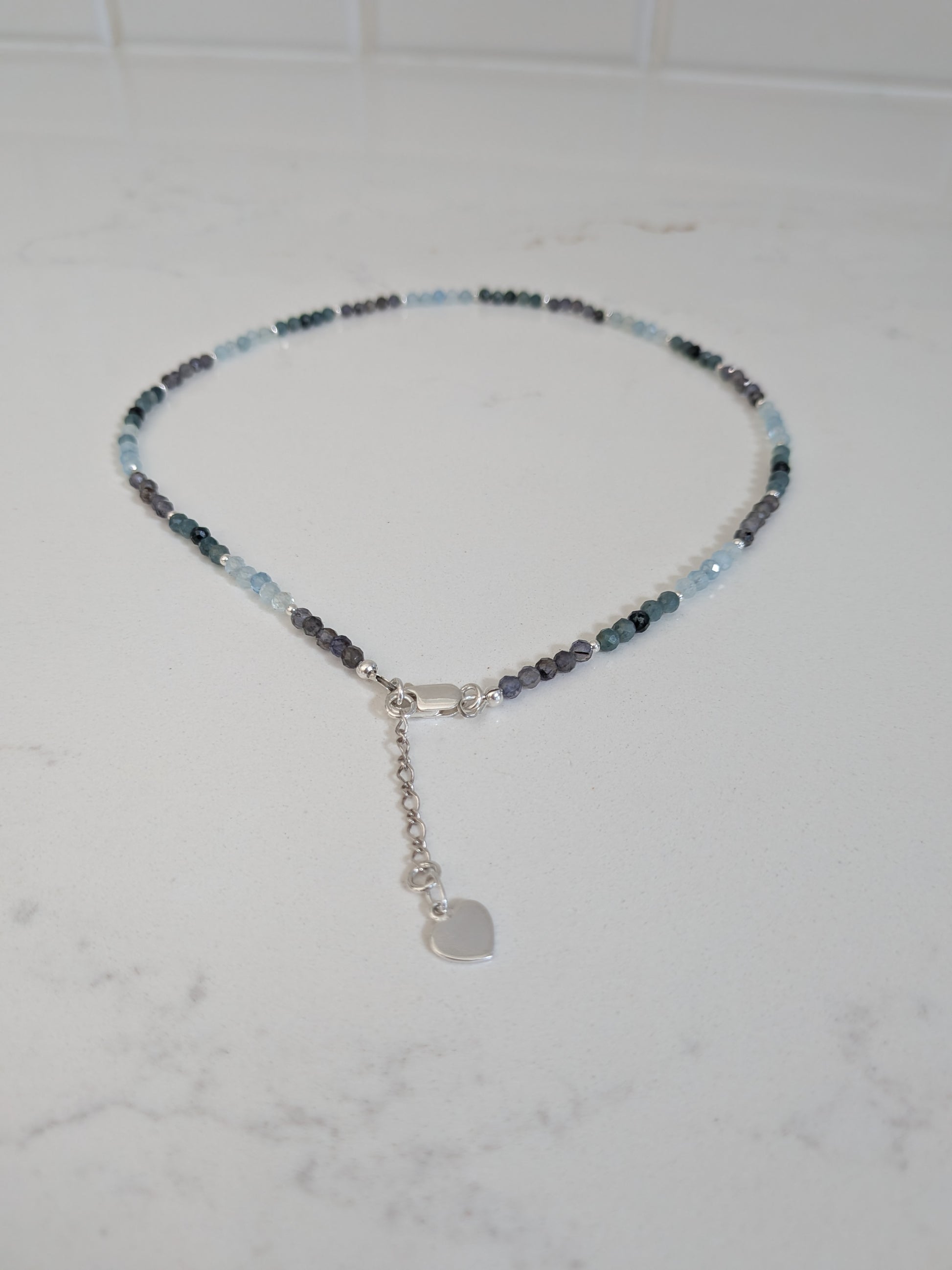 A delicate beaded necklace with small light blue, dark blue, and gray beads, featuring a silver lobster clasp, extender chain, and a small heart-shaped charm, displayed on a white marble surface.