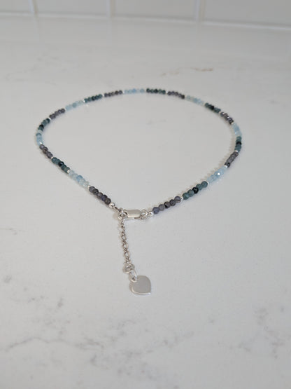 A delicate beaded necklace with small light blue, dark blue, and gray beads, featuring a silver lobster clasp, extender chain, and a small heart-shaped charm, displayed on a white marble surface.