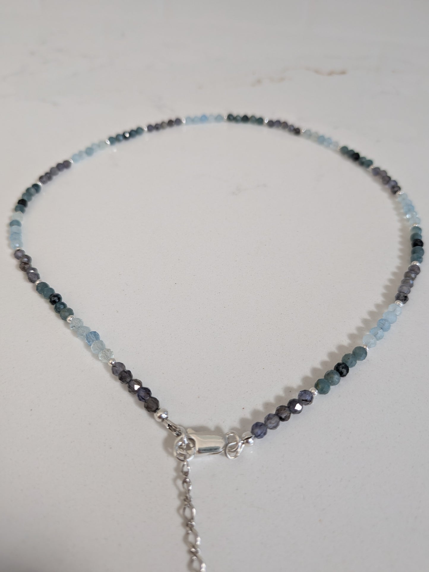 A beaded necklace with faceted light blue, dark gray, and black beads, featuring a silver-toned lobster clasp and extender chain, displayed on a light off-white surface.