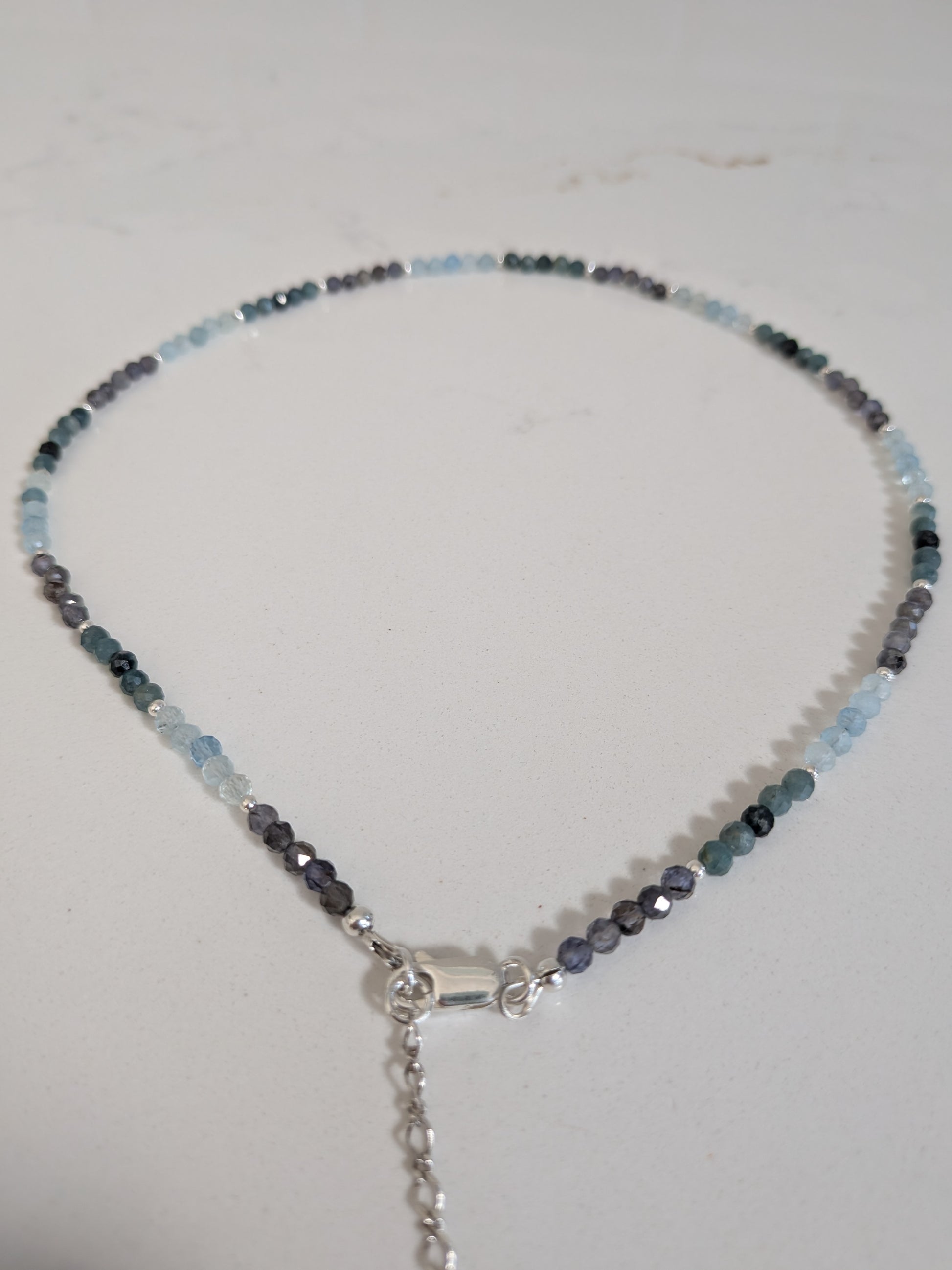 A beaded necklace with faceted light blue, dark gray, and black beads, featuring a silver-toned lobster clasp and extender chain, displayed on a light off-white surface.