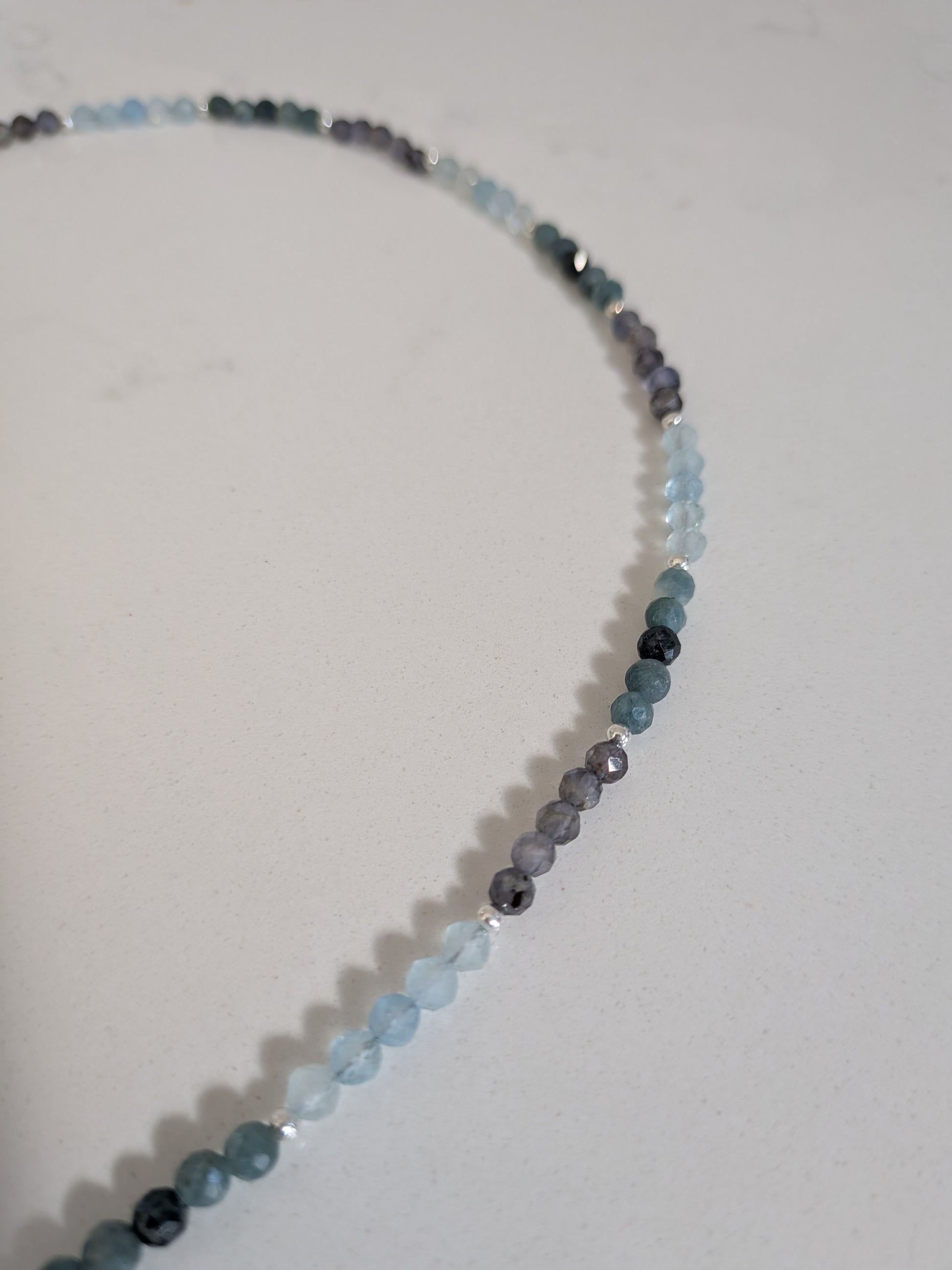 A beaded strand necklace made of small faceted round beads in shades of light blue, medium blue, gray, and dark charcoal, arranged in a curved line on a plain off-white surface.