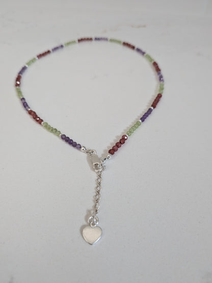 A beaded necklace with small multicolor beads in purple, green, and red, featuring a silver lobster clasp, extension chain, and a dangling silver heart charm, displayed on a light gray surface.